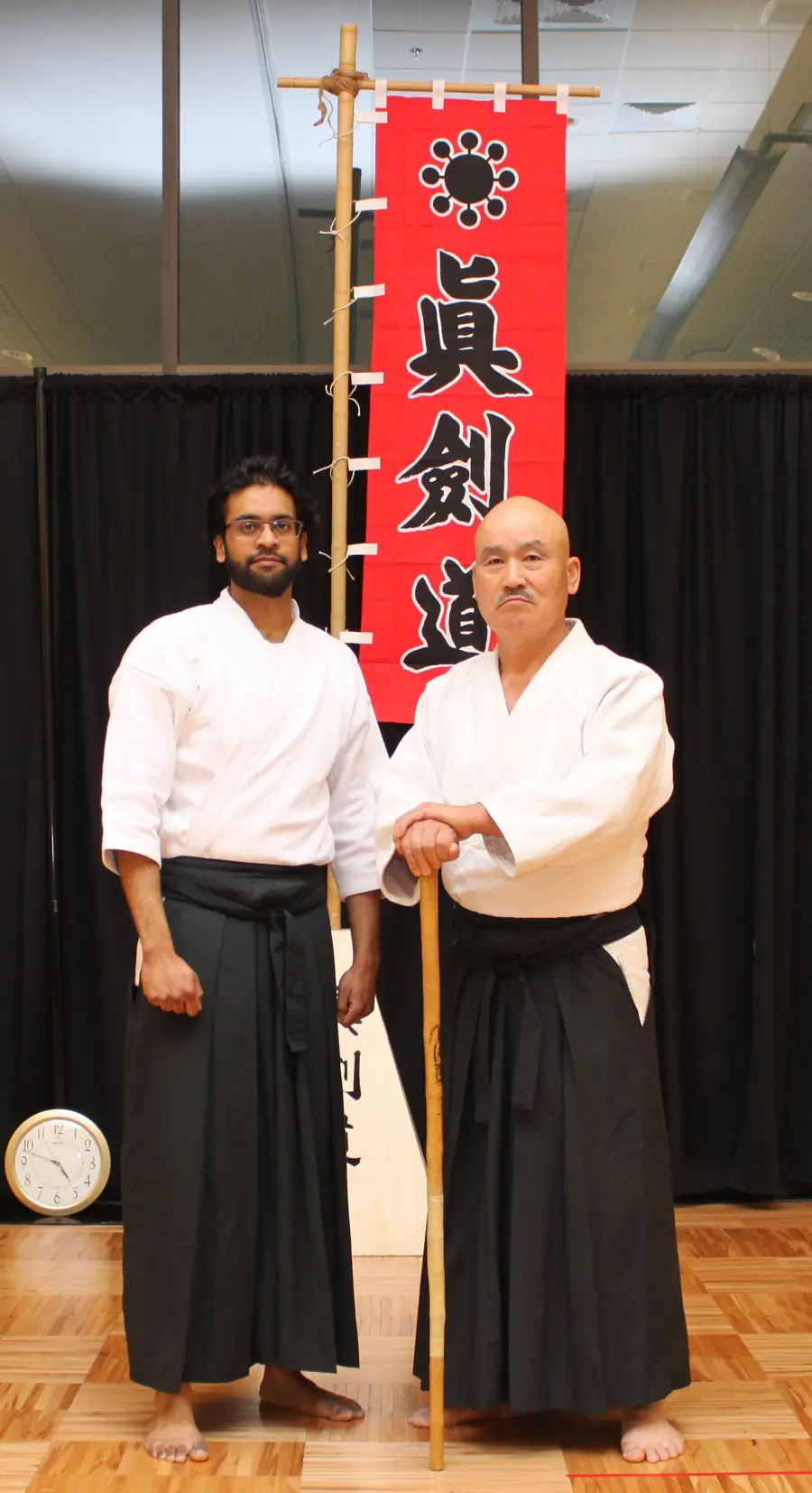 Raheem Syed standing beside Toshishiro Obata Kaiso in front of a red Shinkendo banner