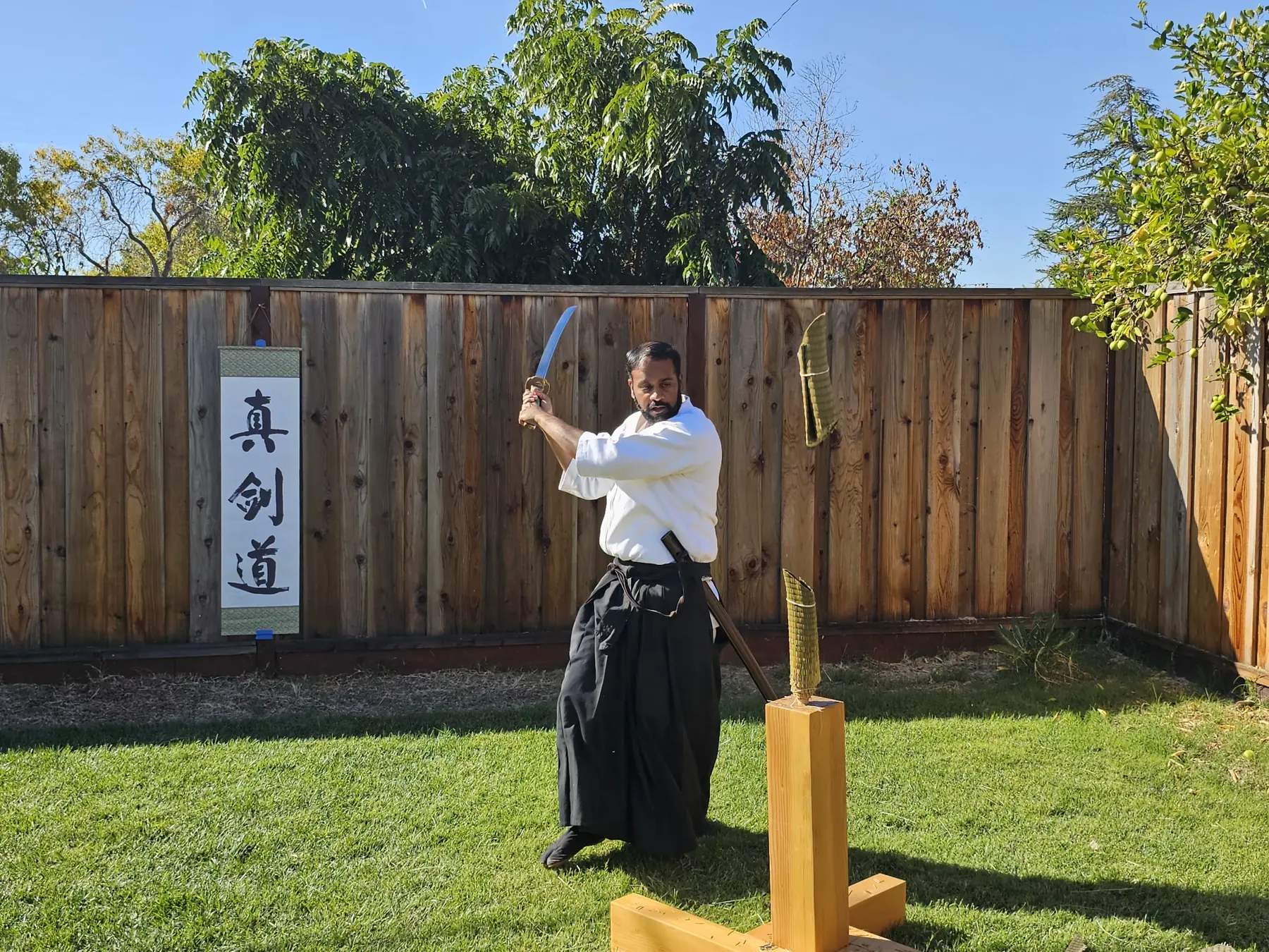 Raheem Syed mid-cut with a live blade at the tameshigiri stand, the Shinkendo scroll beside him